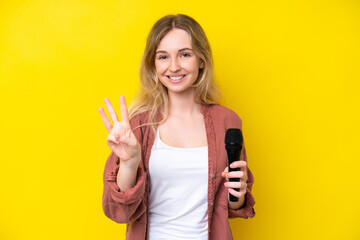 Young singer caucasian woman picking up a microphone isolated on yellow background happy and counting three with fingers