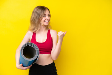 Young sport caucasian woman going to yoga classes while holding a mat isolated on yellow background pointing to the side to present a product