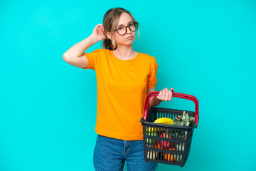 Blonde English young girl holding a shopping basket full of food isolated on blue background having...