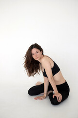 a brunette girl in black leggings and a black short top sits on a white floor against the background of a wall, rests from yoga, dancing or sports exercises, looks at the camera