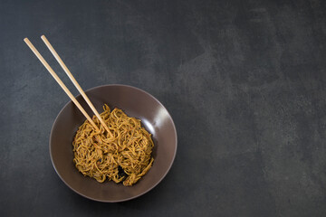 Fideos teriyaki con palillos, en un plato de cerámica marrón, sobre fondo gris oscuro.