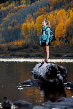 A young woman hiking stands on a log and takes in the view of the fall colors.
