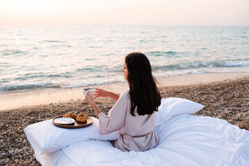 Fototapeta na wymiar Young woman having breakfast in bed with white duvet and pillow outdoor over sea shore at beach. Good morning. Summer vacation season.