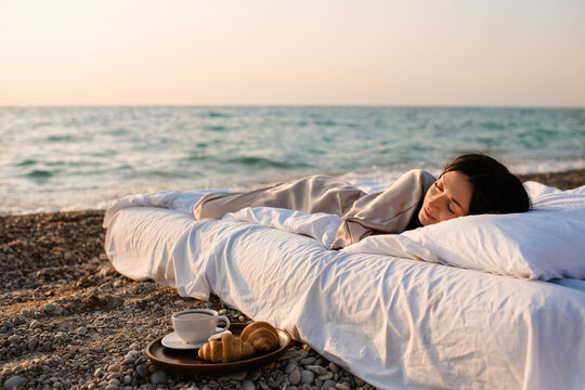 Young woman sleeping in bed on white duvet and pillow with cup of tea with pastry over sea background outdoor. Good morning. Summer vacation season.