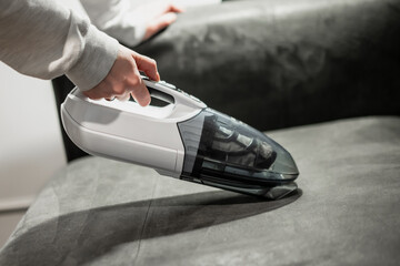Hand held small hand white and grey vacuum cleaner vacuuming a leather couch in grey interior