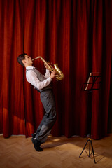 Young musician plays tenor saxophone on stage with red drape curtains - obrazy, fototapety, plakaty