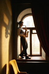 Creative young man playing saxophone standing on windowsill