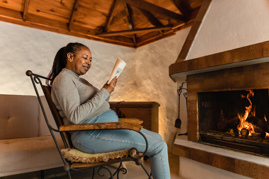 Senior African woman reading book while warming in front of fireplace in her house - Powered by Adobe