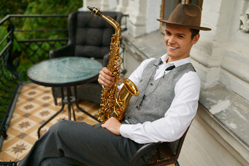 Smiling musician holding saxophone sitting on chair in balcony of his apartment