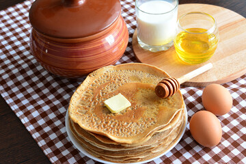 heap of hot fried pancakes on plate on rustic table with tablecloth, honey, ceramic pot and eggs