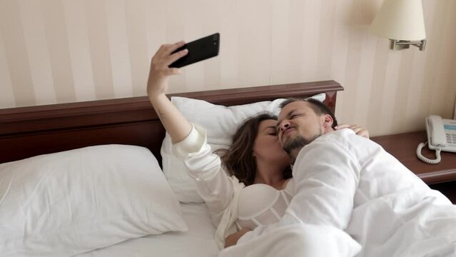 Couple love taking a phone selfie portrait while relaxing in their bedroom together for social media at home. Happy young man and woman smile when taking pictures before posting on online apps