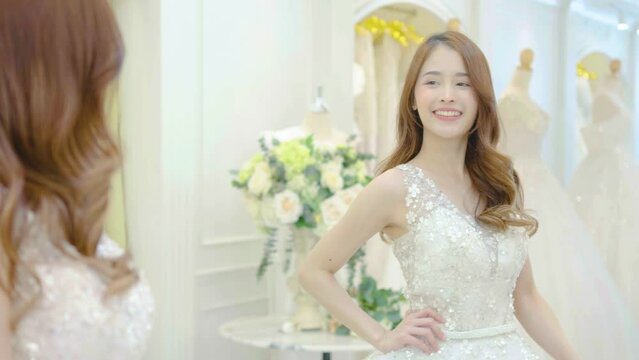 Young asian woman smiling and trying on wedding dress in a shop, asian bride look the mirror for beauty, Young female check beautiful reflection with happy smile for marriage, luxury or fashion