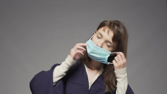 Graduation girl putting on cap and protective face mask. Female student posing against gray background.