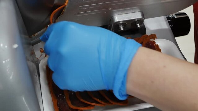 Close-up slicing meat basturma using an electric industrial slicer. Dark red slices of meat in paprika are folded by the seller's hand into a substrate after cutting with a special cutting machine