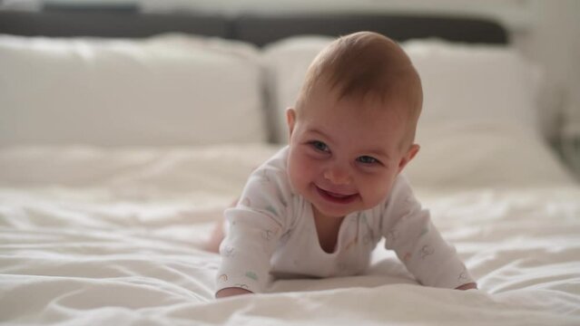 Smiling baby with blue eyes and in white t-shirt lies on belly on bed. Kid puts hands in front and try to hold body. Close-up. First year of life and knowledge of world around. Side view