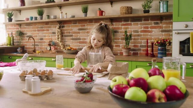 Happy and pretty little girl at the kitchen island using the roller and add some flour over the dough she happy take some juice and drink in front of the camera. 4k