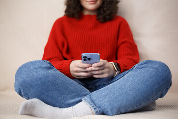 Young girl sitting on couch and typing message on mobile phone. White female person texting online with modern smart phone gadget
