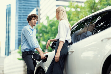 Progressive businessman and businesswoman with electric car connected to charging station before...