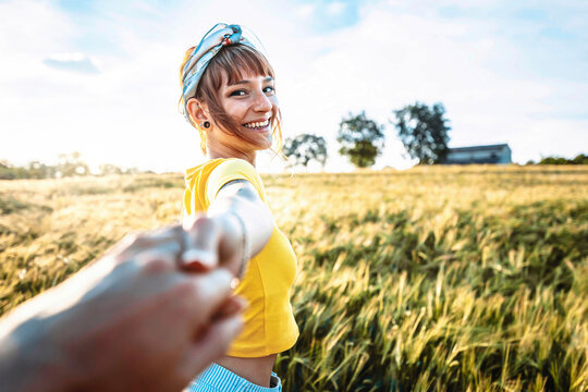 Happy beautiful young woman smiling at camera outside - Loving girlfriend holding boyfriend hands walking on sunset field - Travel, healthy lifestyle, self care, love and wellbeing concept