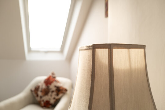 Shallow focus of a fabric bedside lampshade seen in a newly renovated home. A distant skylight window and dressing chair is visible in this airy room image.