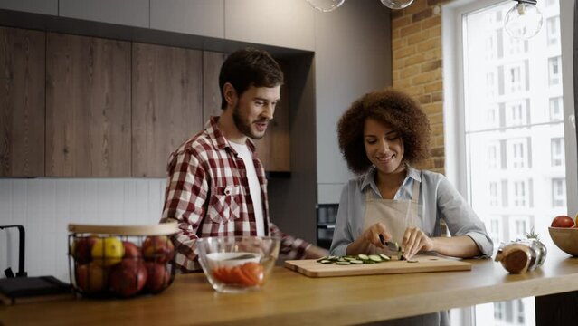 Young man joining his girlfriend who's cooking in the kitchen, couple flirting