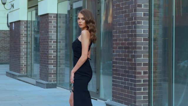 Sexy fashion model woman walking along city street gray sidewalk. Long loose brunette hair fly in wind motion. Wave curl hairstyle. Black evening trendy prom dress. Beauty strong face elegant makeup
