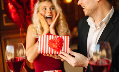 Young couple having a romantic dinner and toasting with glass of red wine. Exchange of gifts. Relationship, surprise and love concept.