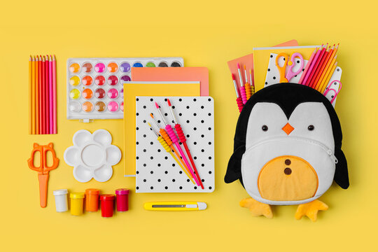 Plush penguin backpack with stationery and supplies for drawing and craft on yellow background. Various colorful material for creativity and art activity. Primary School or kindergarten.
