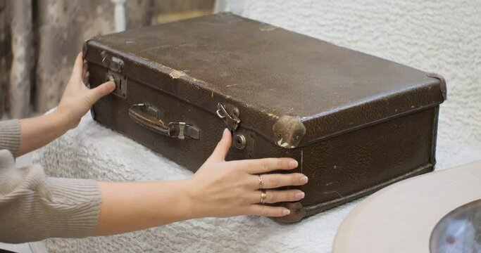 An old brown leather suitcase sits on a white armchair. Women's hands open the locks on the retro suitcase and flip the lid. An old shabby suitcase evokes nostalgia of the past.