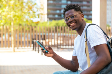 Portrait of young student sitting in the park, using his mobile phone and looking at camera - smile happy man - Education concept
