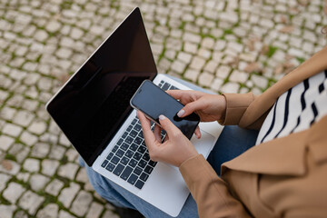 pretty curly woman sitting in city street in stylish jacket, working remote job on laptop