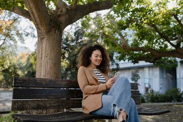 pretty curly smiling woman walking in city street in stylish jacket, using smartphone