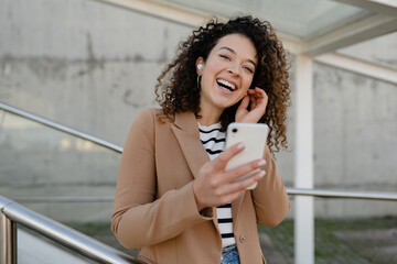 pretty curly smiling woman walking in city street in stylish jacket, talking on smartphone