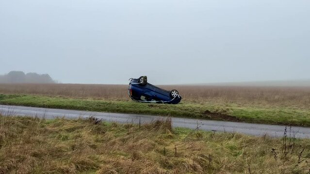 an overturned blue Suzuki hatchback car resting on its roof having crashed off a road on to grass