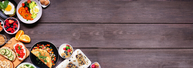 Healthy breakfast food corner border. Overhead view on a dark wood banner background. Table scene with omelette, nutritious bowl, toasts, granola bars, smoothie bowl, yogurts, fruit. Copy space.
