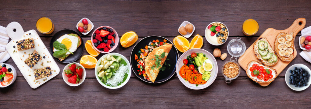 Healthy breakfast food table scene. Top down view on a dark wood banner background. Omelette, nutritious bowl, toasts, granola bars, smoothie bowl, yogurts and fruits.
