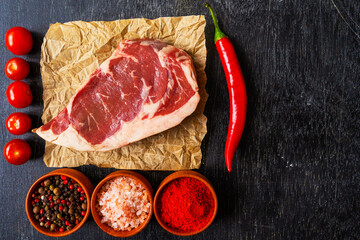 Raw piece of ribeye beef meat on craft paper on a wooden table with three jars of spices, tomatoes, hot peppers ,spice and himalayan salt. Fresh food. Everything for a barbecue.