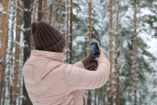 a girl in a pink jacket takes pictures of the winter forest on her smartphone.