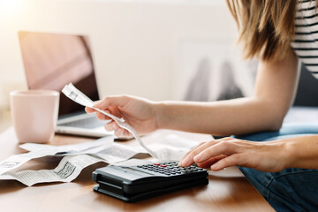 Young homeowner woman sitting on sofa using calculator and laptop to calculate monthly budget, household expenses, mortgage, rental fees, paying bills online