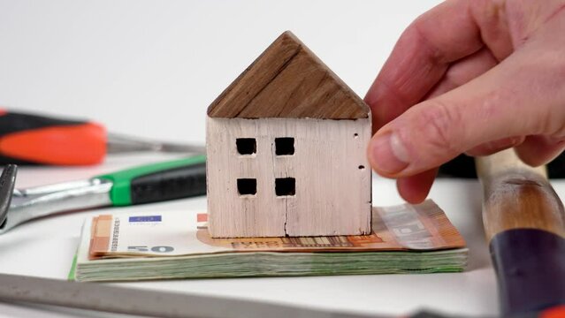 A hand puts a wooden cracked house model on a bundle of money with tools. The concept of a financial project for the renovation and remodeling of a house