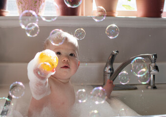 Baby taking bath in the kitchen sink. Child playing with foam and soap bubbles in the sunny...