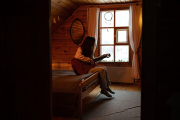 Young woman plays guitar and enjoys music alone, sitting by window of village house with cozy bed,...