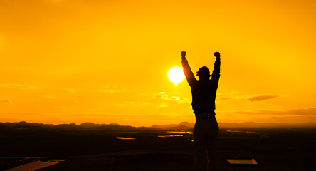 Fototapeta na wymiar Person with arms up as a sign of self-improvement and motivation. Silhouette of a man at sunset on top of a mountain. Warm orange sky. Concept purposes, challenges. Copy space to the left