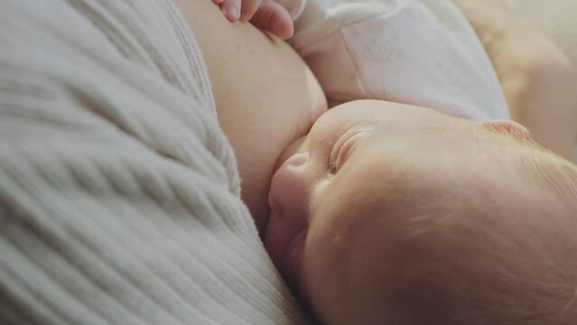 Close-up view of a cute baby with black eyes eating mom's milk. Mother is breastfeeding the baby on the bed