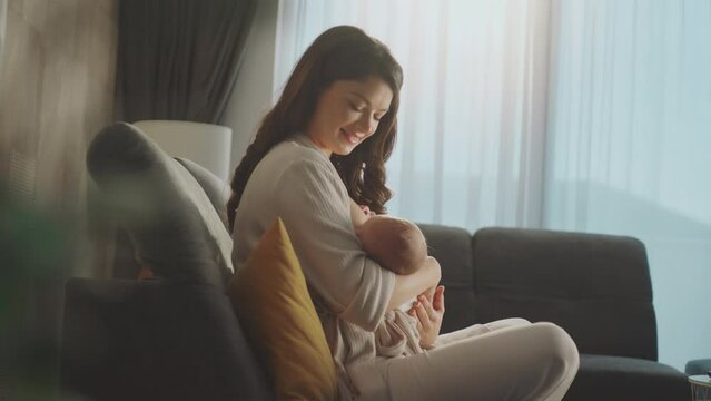 Young mother sitting on sofa and looking at her newborn baby while baby sucking milk from breast. Concept of healthy and natural baby breastfeeding nutrition