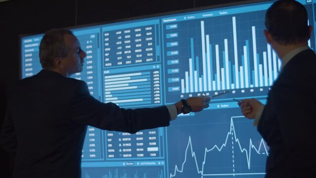 Financial data analysts analyzing charts on big screen. Back view of men in official suits having meeting in modern control center, analyzing rise and fall of prices. Occupation, technology concept