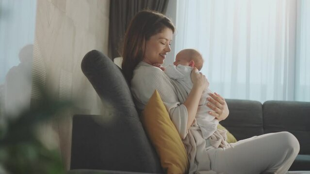 Medium shot of a smiling mother holding baby girl in arms and touching gently in slow-motion while she is sitting on the couch at home