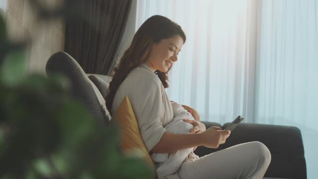 Young mother browsing social media on smart phone when breastfeeding baby at home during daytime. Slow-motion shot