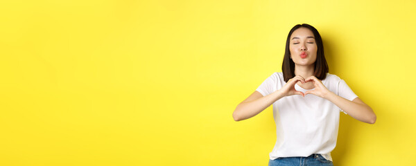 Beautiful asian woman showing I love you heart gesture, smiling at camera, standing against yellow...