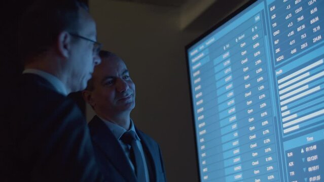 Cheerful businessmen analyzing big data on big digital screen. Financial analysts talking in modern monitoring room, solving work issues, processing information in office. Occupation concept 